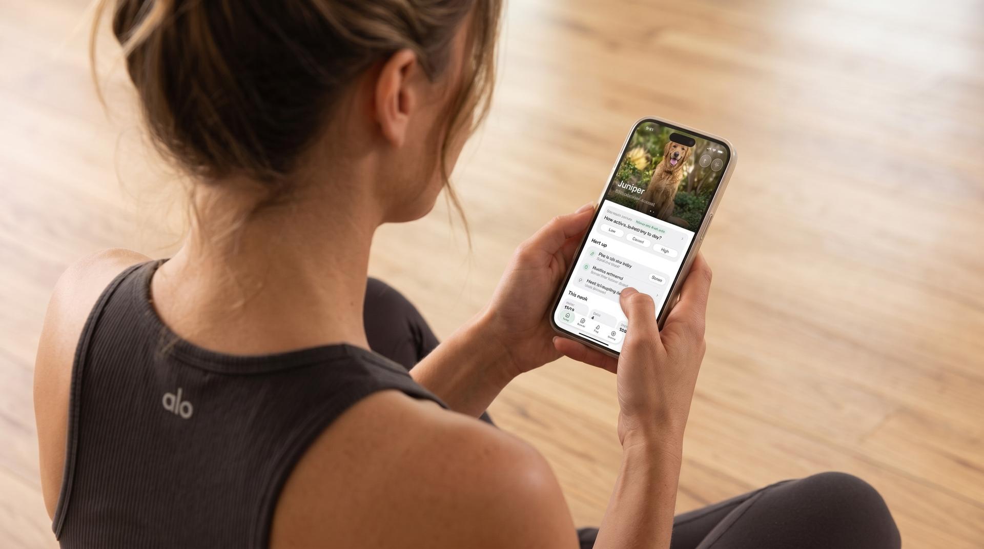 A woman sitting on a wooden floor looks at her dog's profile in the Eva app on her phone
