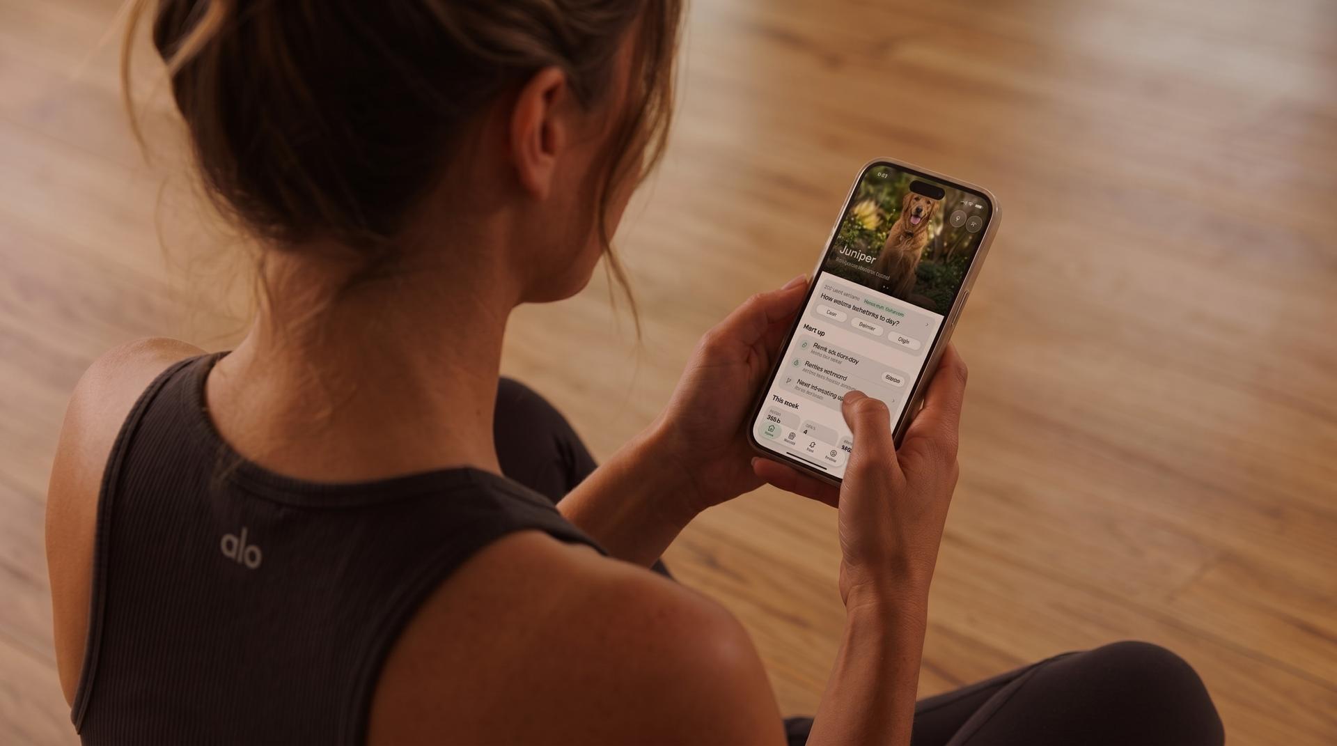 A woman sitting on a wooden floor looks at her dog's profile in the Eva app on her phone